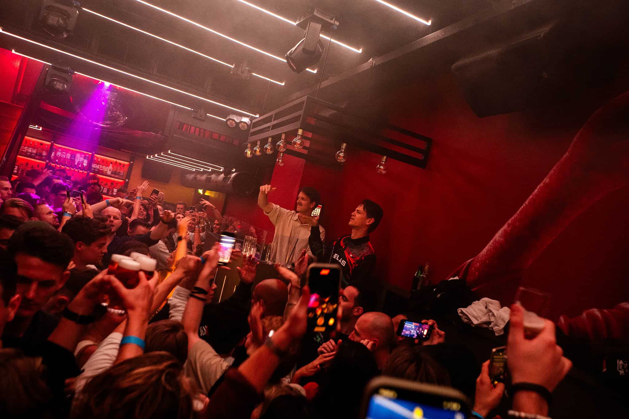 Mensen dansen en feesten in de bar bij ROTO Maastricht, terwijl DJ's Mr. Belt & Wezol hun set draaien en het publiek foto's maakt.